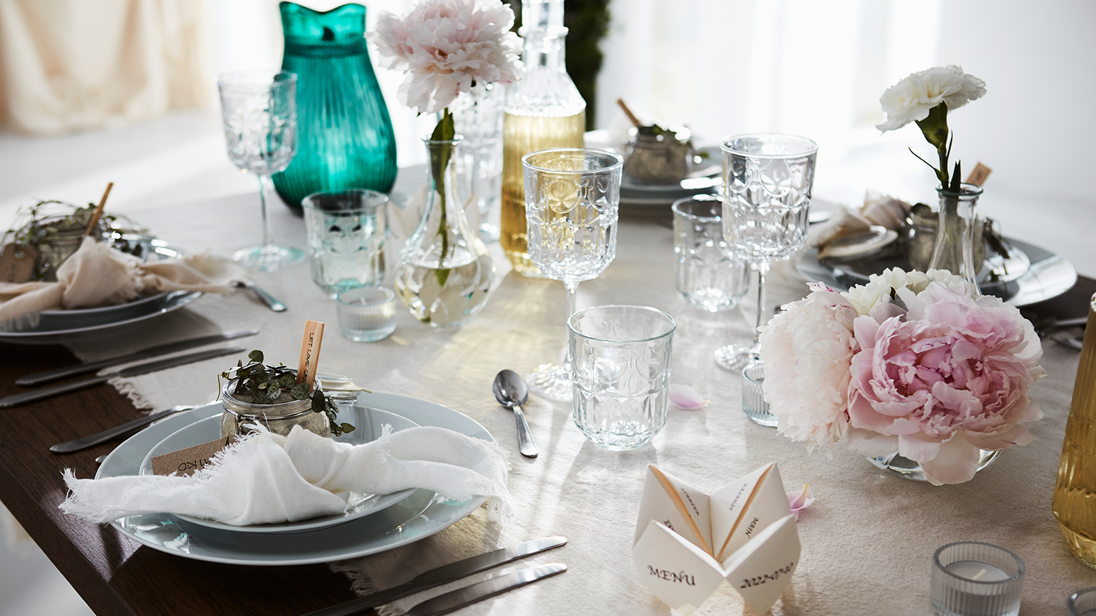 Een gedekte tafel met borden, glazen, bloemen in vazen en verschillende decorstukken. 