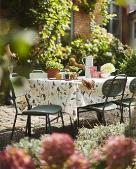 Buitentafel met groene tuinstoelen en vrolijk tafelkleed, in een tuin met veel groen.